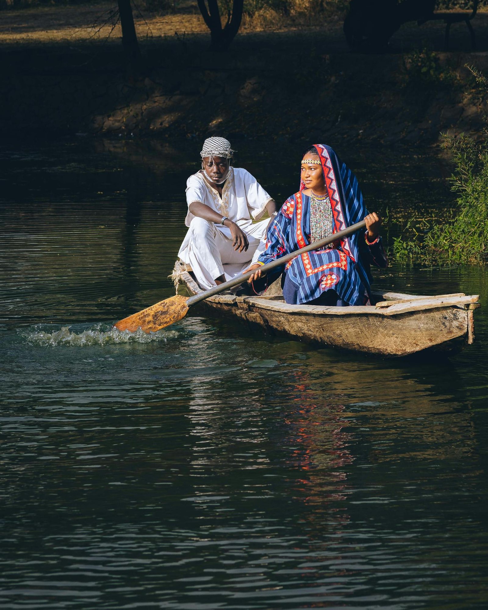 Two Nigerian adults in traditional clothing paddle a wooden canoe on a serene river.